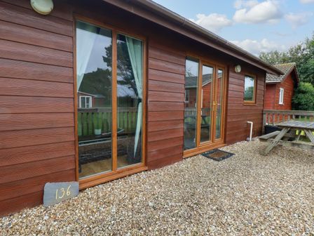 A wooden cabin exterior with glass doors and windows and a picnic table on gravel at Gwanda Tregolds near St Merryn
