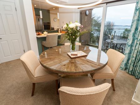 A dining area with a round wooden table and four chairs next to a kitchen and a balcony with a view at Fal View in Falmouth