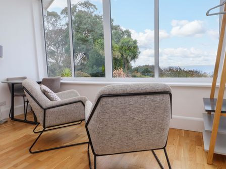 A sitting area with two armchairs a small table and large windows showing trees outside at Summer Room in Llanbedrog