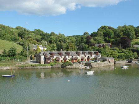 A row of waterfront houses with boats on a river and trees on hills behind at Gillyweed in Noss Mayo