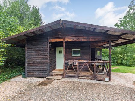 A wooden log cabin with a small porch and outdoor chairs surrounded by trees at The Log Cabin in Sheldon Blackdown Hills
