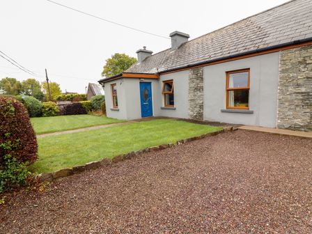 A house with a blue door and two windows with stone and gray walls with a gravel driveway and green grass at Susie's Cottage in Shanacloon near Beaufort County Kerry