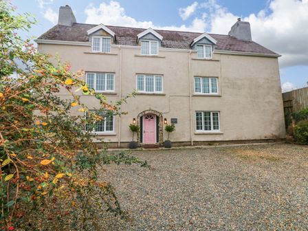 A beige house with a pink door and white windows with shutters surrounded by gravel and some shrubs at Norchard Farm House Redberth near St Florence