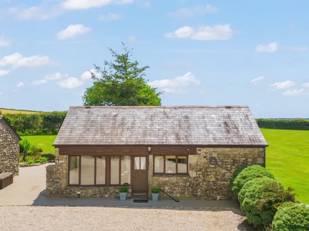 A stone cottage with a shingled roof and a brown door surrounded by green bushes and grass at The Dairy in St Neot