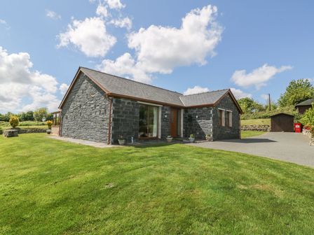 A stone house with a grassy lawn and a paved driveway under a blue sky at Bwthyn Clyd in Llanddaniel Fab