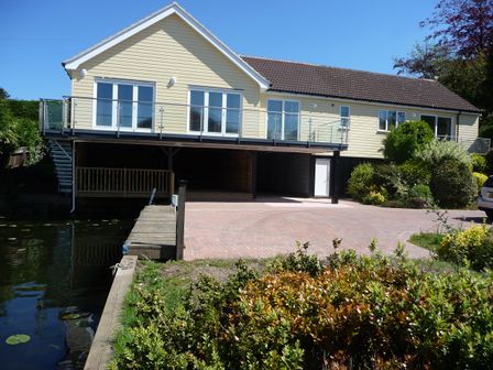 A raised house with a balcony over a carport and a driveway next to a water body with plants at Reedlings in Horning