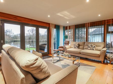 A living room with beige sofas a glass coffee table and large windows overlooking water at Tealby in Horning