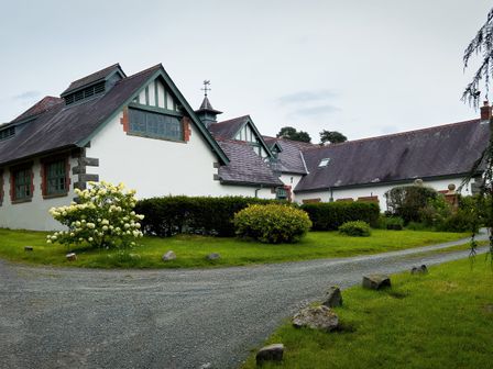 A driveway bordered by grass leading to white buildings with pitched roofs and greenery around at Coach House in Llanwrtyd Wells