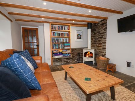A living room with a brown sofa and blue cushions a wooden coffee table a bookshelf a wood burning stove and a wall mounted tv at Crab Cottage in Delabole