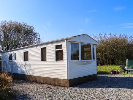 A white mobile home on a gravel surface with trees and outdoor chairs and plants nearby at Hayesland Holiday Home Kilrane near Rosslare Harbour County Wexford