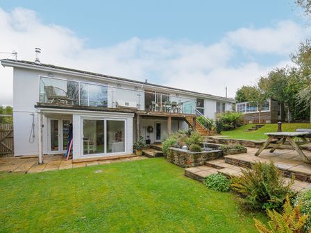 A backyard with a grassy lawn a wooden deck with glass railing patio furniture and a stone planter at Seamay in Polzeath