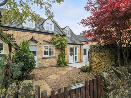 A stone cottage with white windows and doors surrounded by trees and a small garden at 4 Noel Court in Chipping Campden