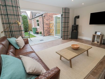 A living room with a brown leather sofa, wooden coffee table, wall-mounted television, and open doors leading to a patio at Breamish Christon Bank near Embleton