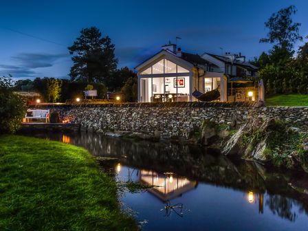 A modern house with large windows lit up next to a stone wall and a small river at River Barn in Town End