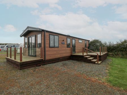 A wooden cabin with glass railings on a raised deck with outdoor furniture surrounded by gravel and grass at Caldey Retreat in Hasguard Cross near Broad Haven
