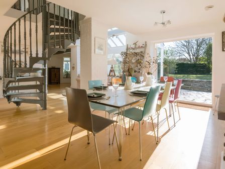 A dining area with a table set for six, multicolored chairs, spiral staircase, and glass doors opening to a garden at Moorlands Bungalow in Rock