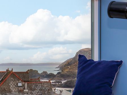 A coastal view with rocky cliffs and rooftops seen through a window with a blue cushion inside Mavericks Wave in St Agnes