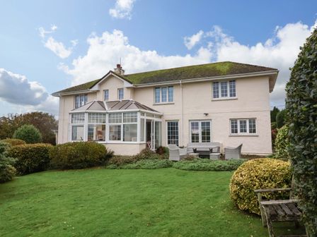 A two-story house with a conservatory and garden furniture on a lawn surrounded by bushes at Heanton Lodge in Heanton near Braunton