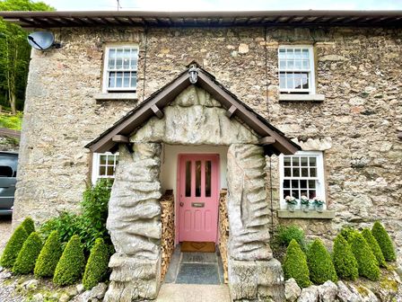 The stone exterior of a house with a pink front door and small shrubs at Pear Tree Cottage in Grange-Over-Sands