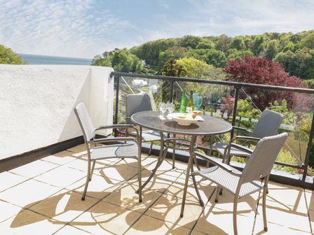 A balcony with a round table and four chairs overlooking trees and the sea at Valley View 9 St Elmo Court in Salcombe
