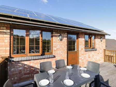 An outdoor dining area with a table set with plates glasses and cutlery on a wooden deck outside a brick building with solar panels on the roof at Avon View in Welford-On-Avon