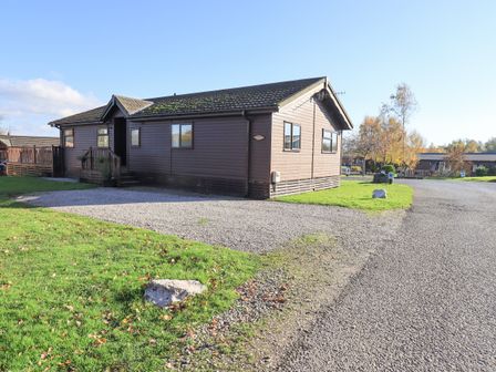 A brown wooden lodge with a gravel driveway and green grass at Kellet Lodge Lakeside 26 Dock Acres near Carnforth