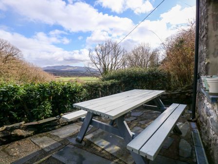 An outdoor seating area with a wooden picnic table and benches surrounded by bushes and trees with a view of distant hills at Osprey Cottage in Prenteg near Porthmadog