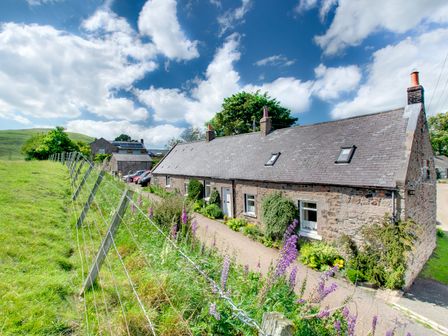 A stone cottage with a gray roof and flower garden next to a fenced grassy field at Braeside High Humbleton near Wooler