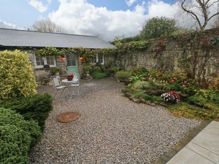 An outdoor courtyard with gravel ground a small metal table and two chairs stone walls and a small garden with plants and flowers at Little Cloud in Nash near Cowbridge