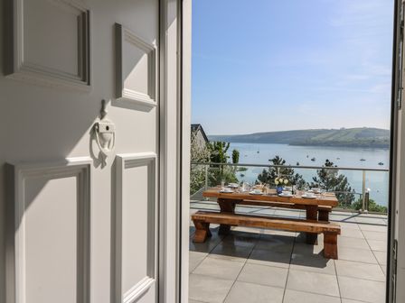A wooden picnic table set with dishes and glasses on a tiled balcony overlooking a body of water with boats and hills in the background at Fronwig in New Quay