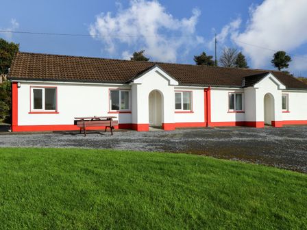 A white house with red trim and brown tiled roof with a wooden picnic table in front and green grass in the foreground at Robin's Roost 2 in Cornamona County Galway