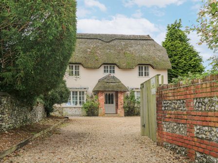A gravel driveway leading to a thatched roof cottage with brick and flint walls and surrounding trees at Moles Cottage in Farnham near Sixpenny Handley