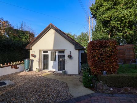 A small white building with a door and windows surrounded by gravel and a garden area at Studio Apartment in Minehead