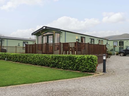 A green static caravan with wooden deck railing surrounded by gravel driveway and green hedges at Highlands Way Lodge in Lamplugh near Cleator Moor