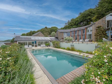 An outdoor swimming pool surrounded by plants and a paved patio with a stone house and glass windows in the background at Curlew House in Cosheston