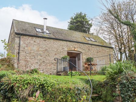 An exterior view of a stone barn with a slate roof and two skylights surrounded by greenery and trees at The Barn at Yeoland House in Yelverton
