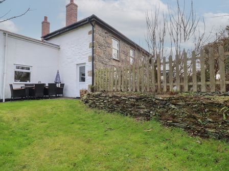 A grassy garden with a stone wall and wooden fence next to a stone and white cottage with outdoor dining furniture at Bramble Cottage in Gwithian near Connor Downs