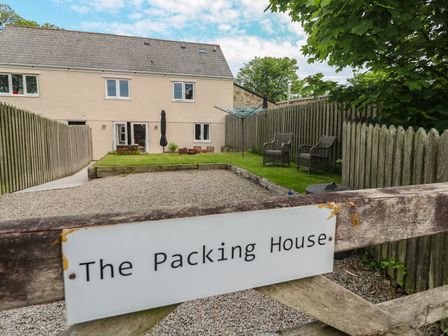 A yard with gravel driveway and lawn with outdoor chairs behind a wooden gate with a sign The Packing House in Rosudgeon