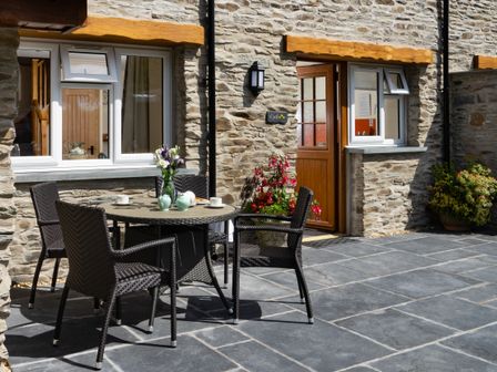 An outdoor patio with a round table set with teacups and vase of flowers and four chairs outside a stone house at Cych Cottage Cardigan Near Cenarth