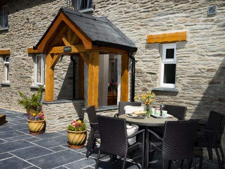 An outdoor patio with a round table and four chairs next to a stone building with wooden porch beams and potted flowers at Towy Cottage Cardigan Near Cenarth