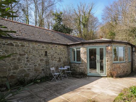 An outdoor stone patio with two white chairs and a small table in front of a stone building with blue framed windows and doors at Rose Cottage in Callington