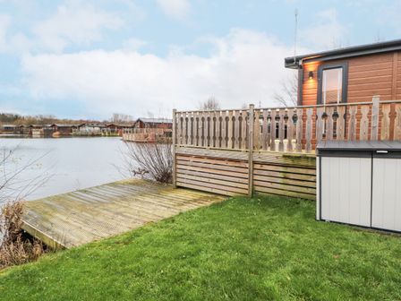 A wooden deck and fenced patio beside a lake with several cabins across the water at 12A Lake Escape in Carnforth