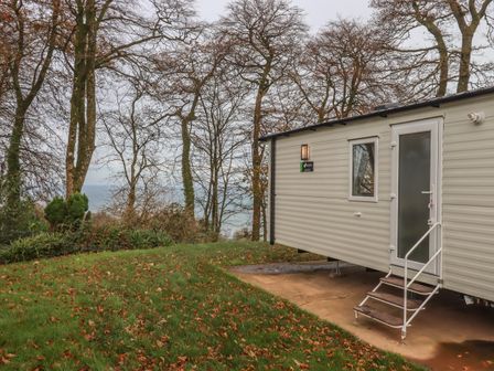 A static caravan with stairs on a concrete pad next to grass and trees with a view of the sea at 5 Manor Park in Bucks Cross Bideford