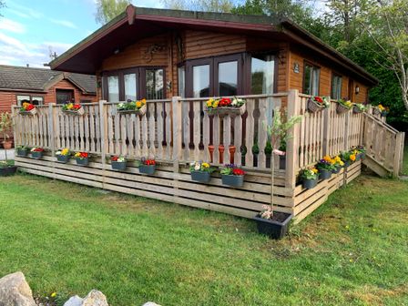 A wooden lodge with a fenced porch decorated with flower pots and a grassy lawn at Lavender Lodge in South Lakeland Leisure Village