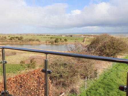A balcony with glass and metal railing overlooking a pond trees fields and the sea at Dunstanburgh View in Embleton