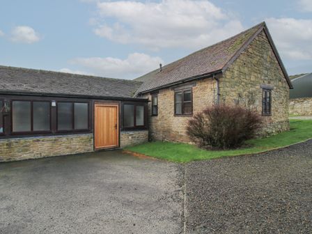 An exterior view of a stone house with a wooden door and windows surrounded by a gravel and paved driveway and green lawn at Walton Maisonette in Craven Arms