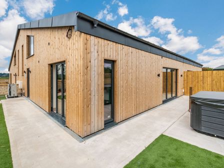 Exterior view of a modern wooden building with sliding glass doors and a hot tub on a concrete patio