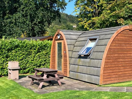 A wooden pod with a small window and door on a stone patio with a wooden picnic table and a concrete barbecue near green bushes at Dovestone Pod 23 in Greenfield