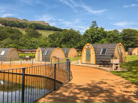 A group of small wooden pods with curved roofs near a fenced pond in a green outdoor setting at Dovestone Pod 18 in Greenfield