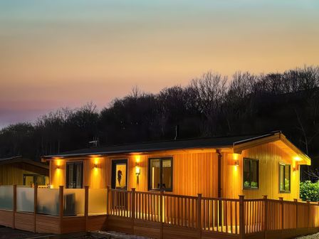 A wooden lodge with illuminated exterior lights and a fenced porch at 12 Green Clough Lodge in Greenfield